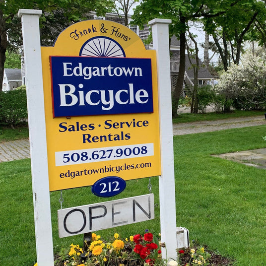Edgartown Bicycle sign with contact information and 'Open' status on a grassy area with flowers.